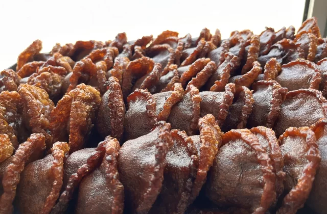 Numerous dark brown cucur cakes with golden-brown ruffled edges are closely arranged on a surface, with a bright, blurred background.