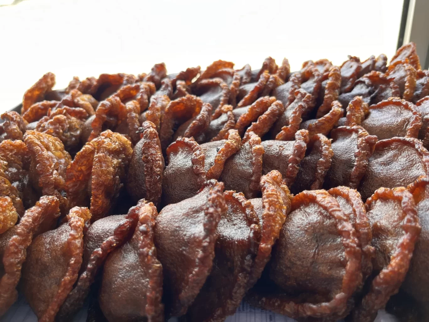 Numerous dark brown cucur cakes with golden-brown ruffled edges are closely arranged on a surface, with a bright, blurred background.