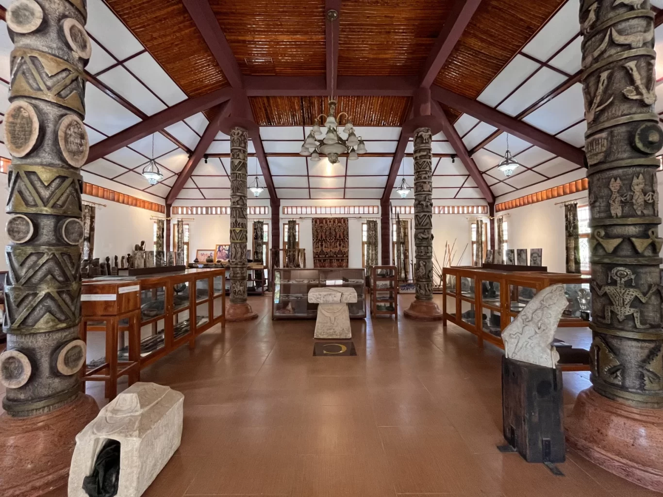 Inside Rumah Budaya Sumba, tall carved pillars frame a hall with display cases, stone artifacts, and a high wooden ceiling.