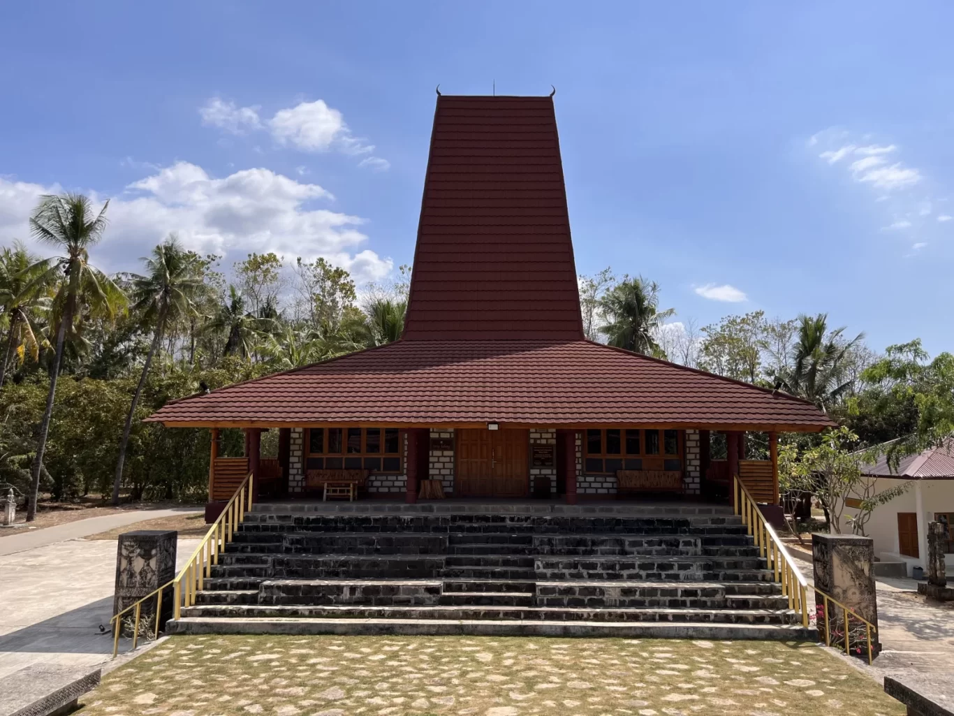 A traditional Sumba-style building with a tall, pointed brown roof and tiled lower roof stands elevated on stone steps, surrounded by green palm trees under a blue sky.