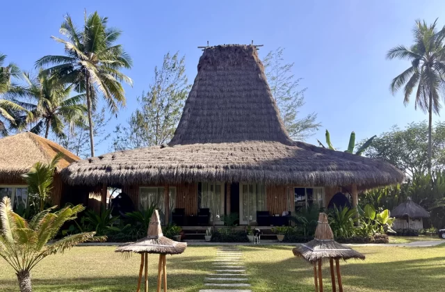 A traditional Sumbanese villa with a tall, conical thatched roof stands on a green lawn under a clear blue sky, flanked by palm trees.