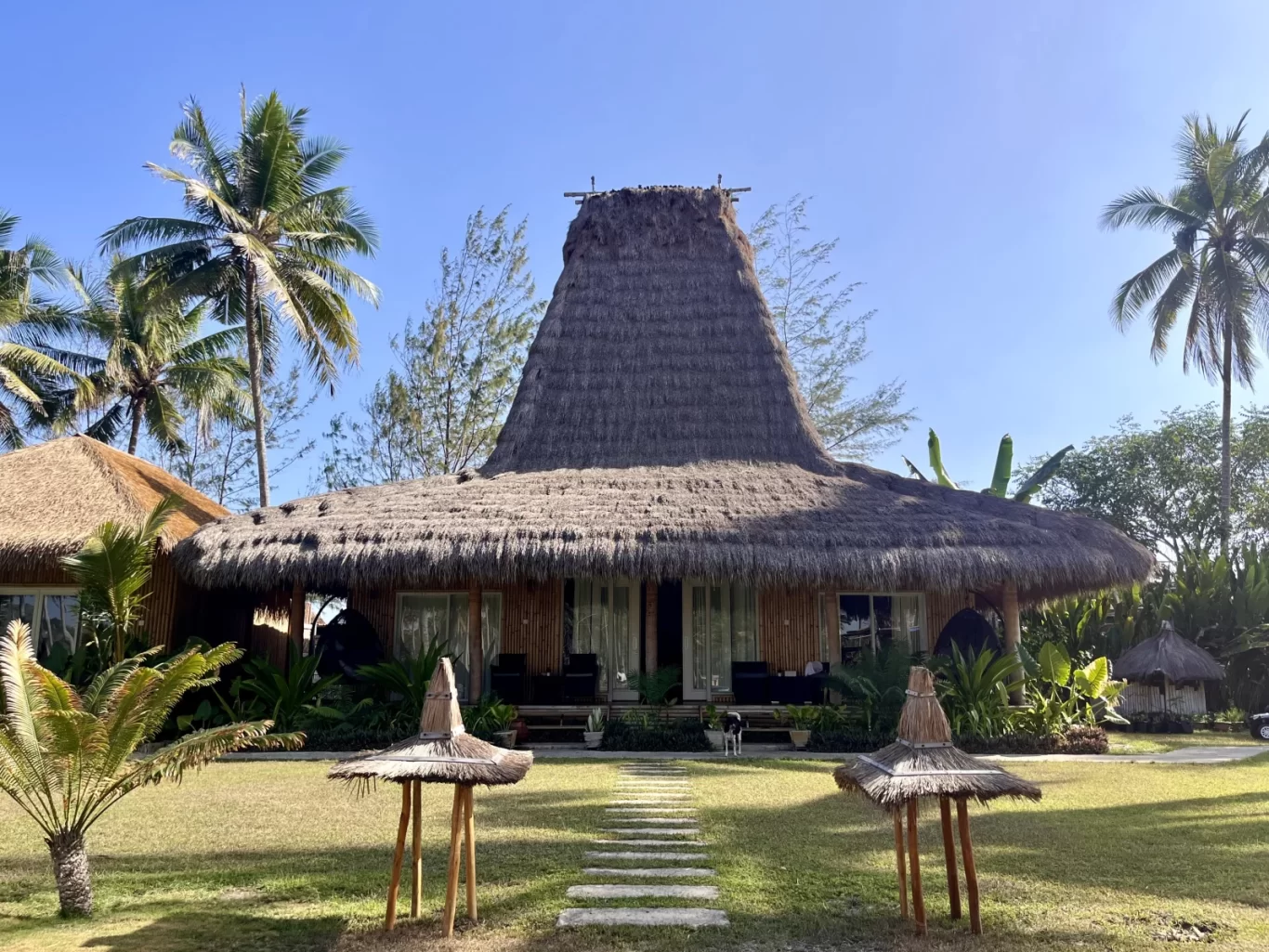 A traditional Sumbanese villa with a tall, conical thatched roof stands on a green lawn under a clear blue sky, flanked by palm trees.