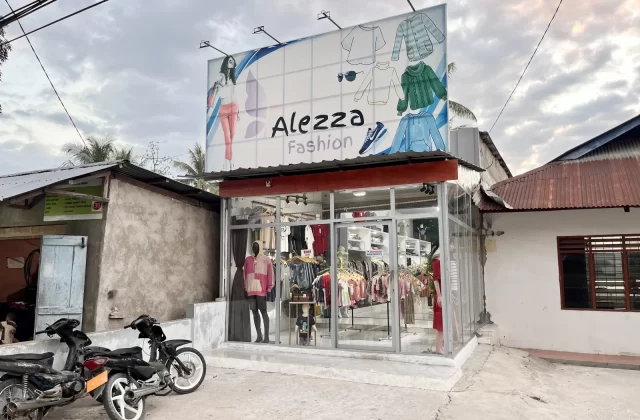 A modern glass-fronted fashion store, Alezza Fashion, stands under a cloudy sky with a large billboard above, two motorcycles parked in front.