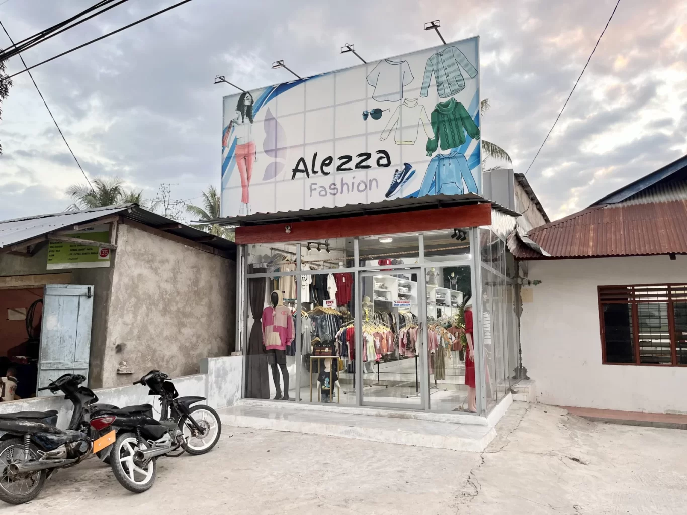 A modern glass-fronted fashion store, Alezza Fashion, stands under a cloudy sky with a large billboard above, two motorcycles parked in front.