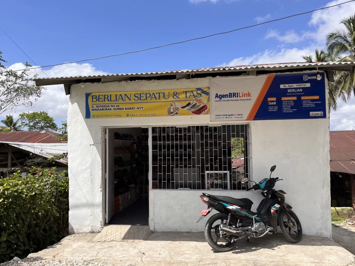 A white building housing Berlian Sepatu & Tas, a shoe and bag store, stands under a blue sky with a parked black motorcycle in front.
