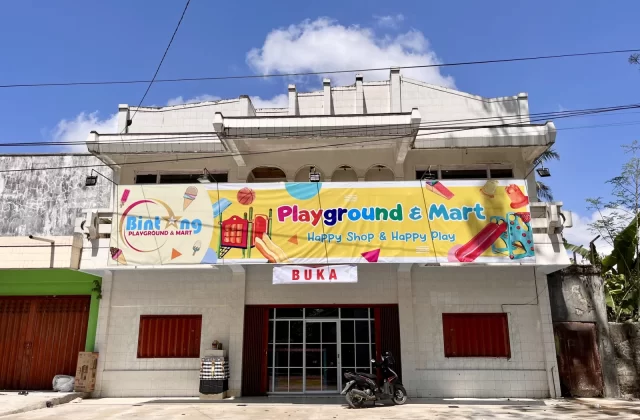 A two-story white building features a large, colorful "Bintang Playground & Mart" banner, with a motorcycle parked in front of its glass entrance under a blue sky.