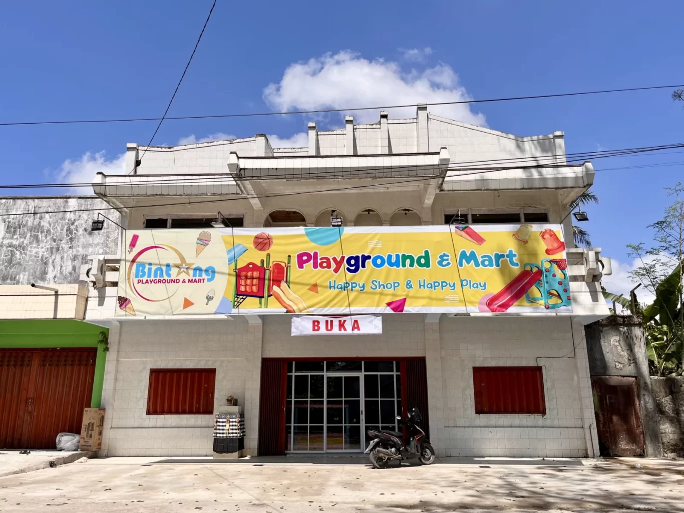 A two-story white building features a large, colorful "Bintang Playground & Mart" banner, with a motorcycle parked in front of its glass entrance under a blue sky.