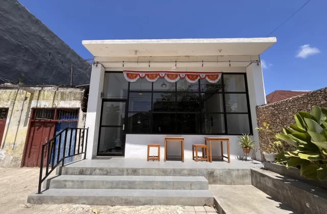 The modern white exterior of Dapur Ma'er restaurant features large glass windows, concrete steps, and a red and white banner under a clear blue sky.