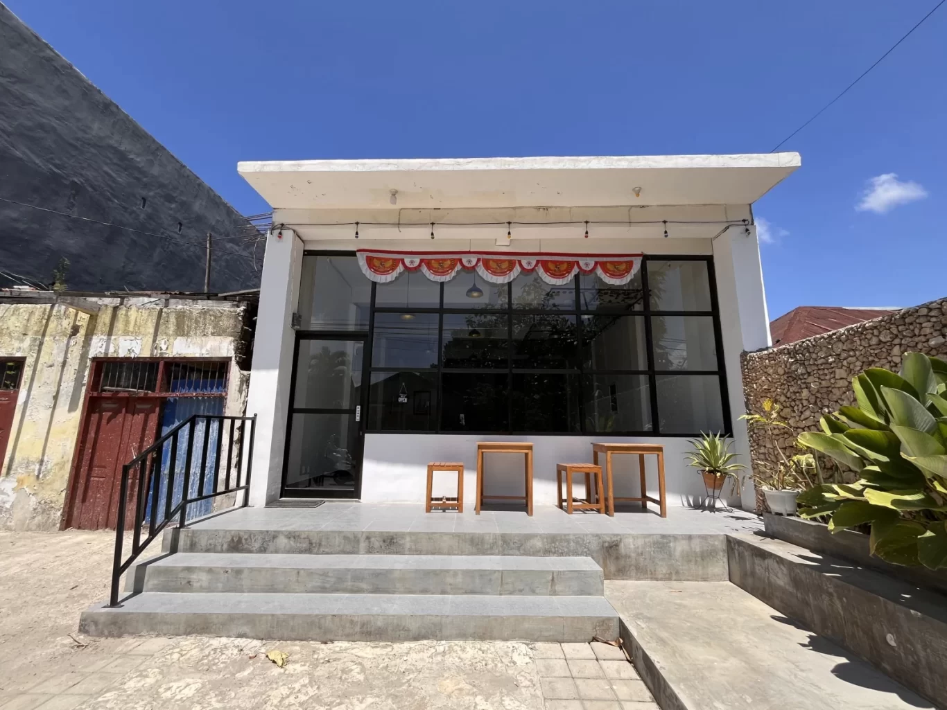 The modern white exterior of Dapur Ma'er restaurant features large glass windows, concrete steps, and a red and white banner under a clear blue sky.