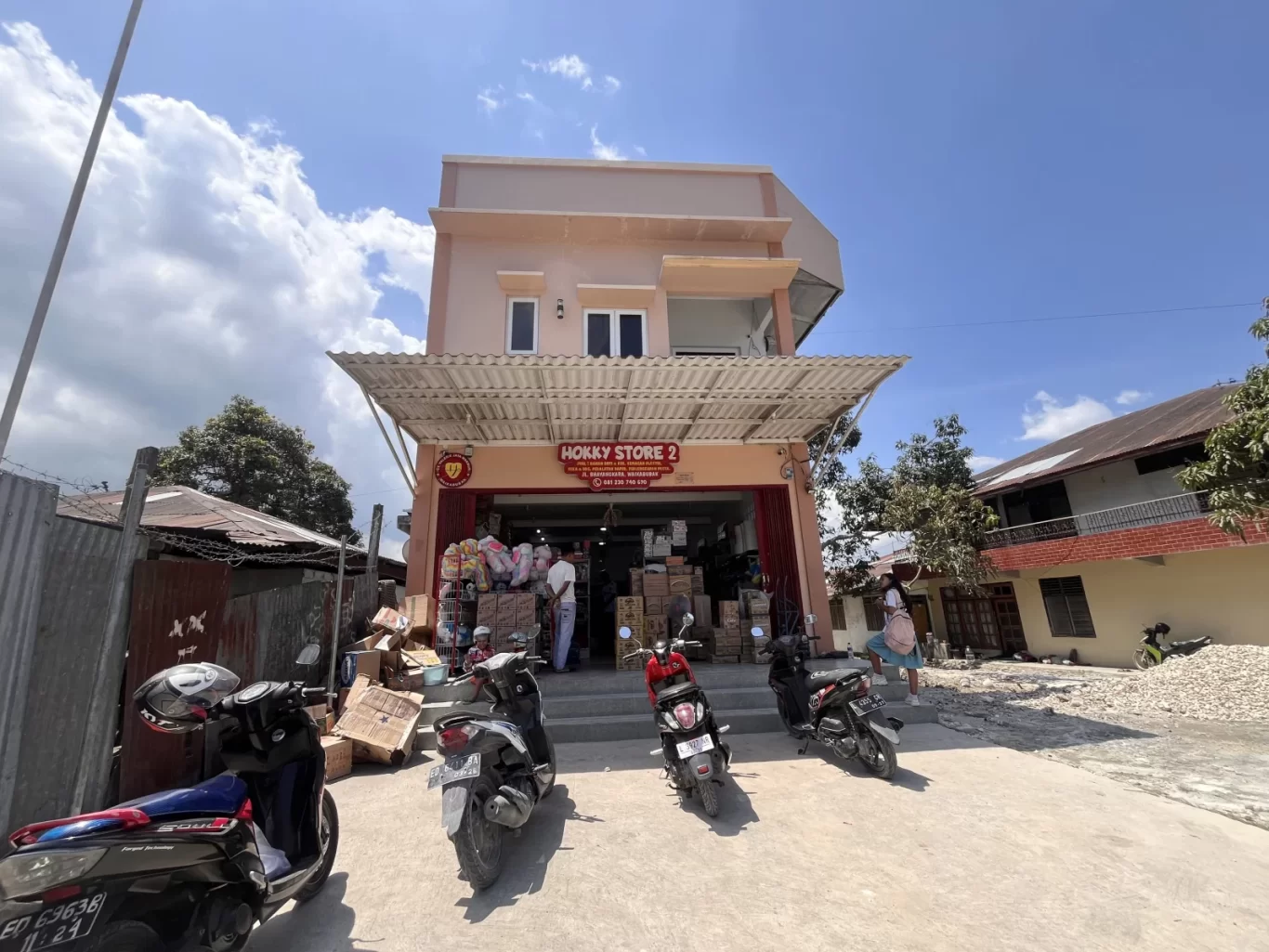Hokky Store 2, a peach-colored two-story building with an open storefront, stands under a blue sky with white clouds, surrounded by parked motorbikes and people in Waikabubak.