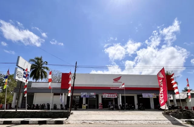 A white and red Honda motorcycle dealership in Waikabubak, West Sumba, under a blue sky, with parked motorcycles and red and white flags.