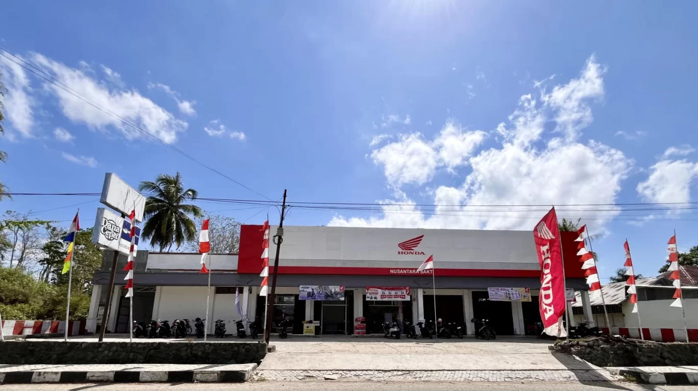 A white and red Honda motorcycle dealership in Waikabubak, West Sumba, under a blue sky, with parked motorcycles and red and white flags.