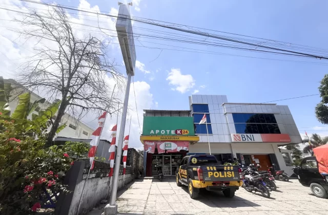The K-24 pharmacy and BNI bank in Waikabubak, West Sumba, stand under a bright blue sky, with a police truck parked outside.