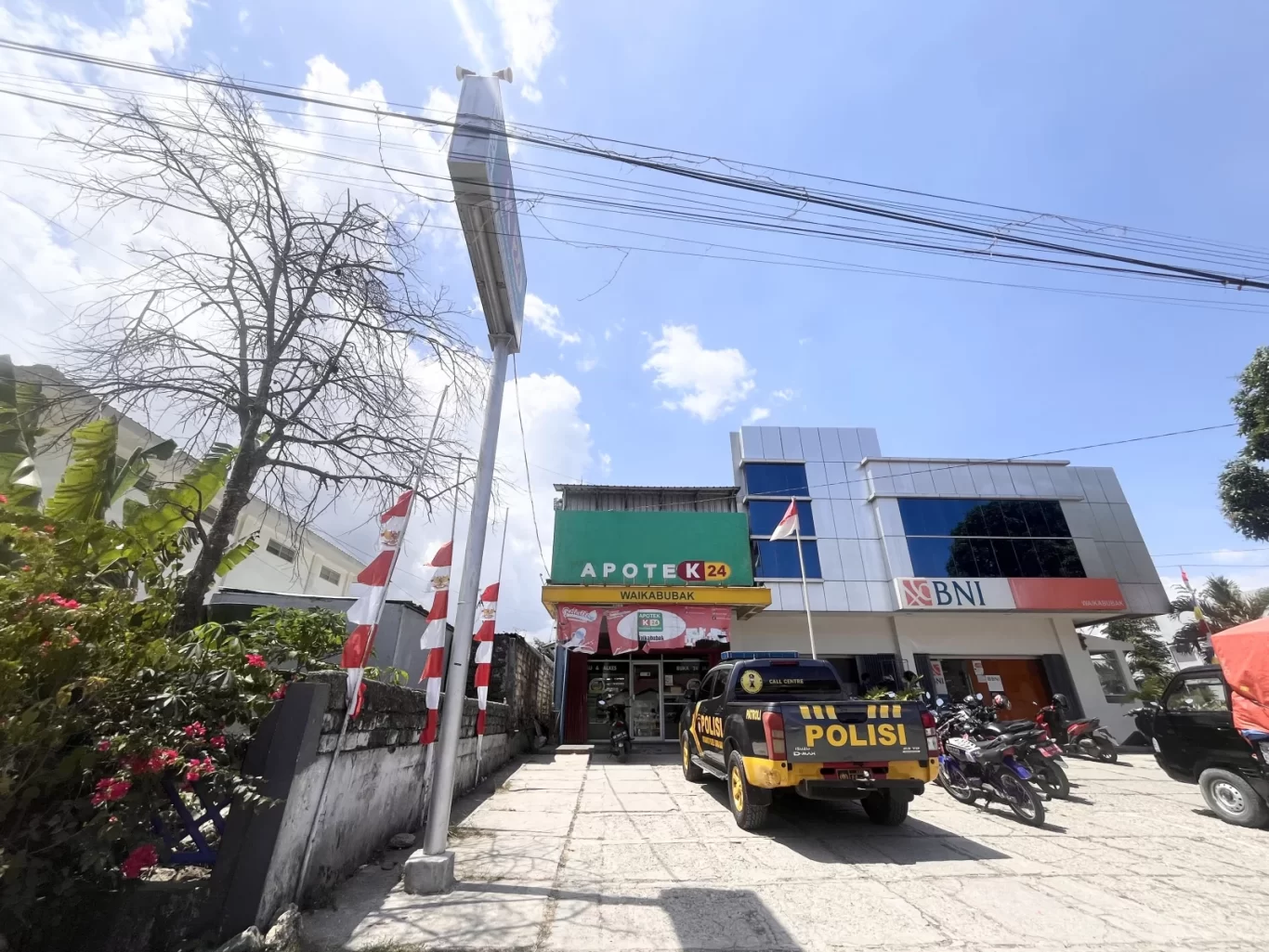 The K-24 pharmacy and BNI bank in Waikabubak, West Sumba, stand under a bright blue sky, with a police truck parked outside.