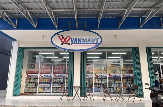 The storefront of Kalena Wano Winmart in Tambolaka features a blue and white facade, large glass windows displaying products, and outdoor seating.