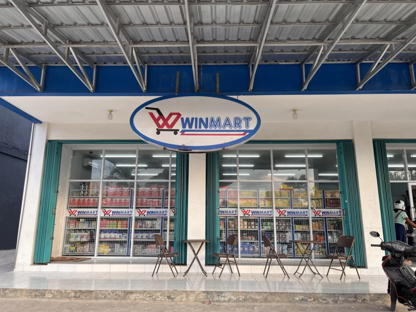 The storefront of Kalena Wano Winmart in Tambolaka features a blue and white facade, large glass windows displaying products, and outdoor seating.