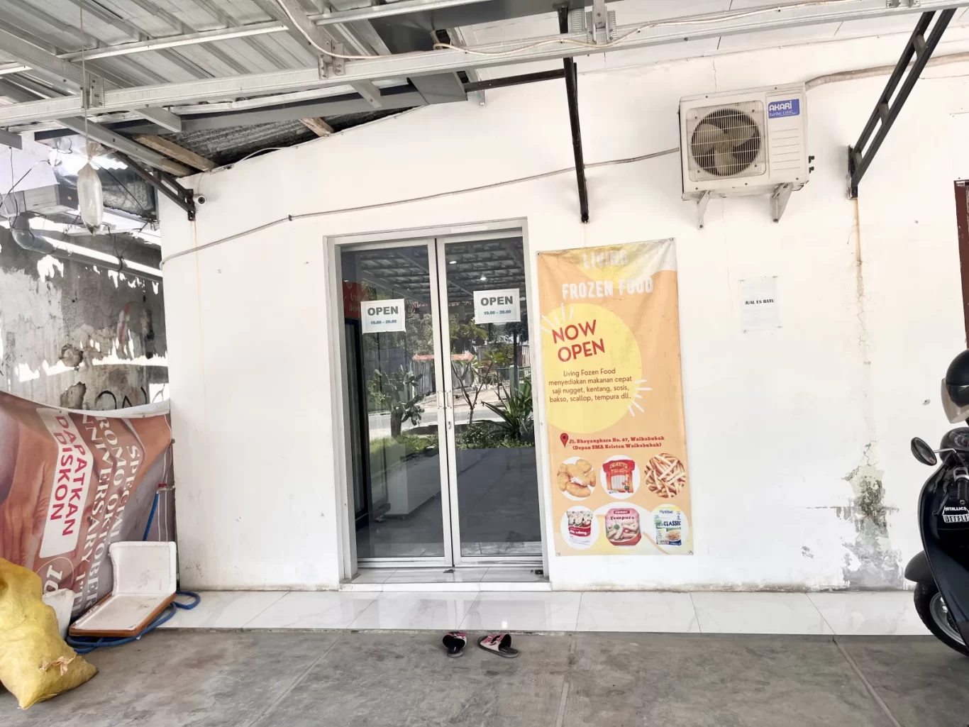 The white exterior of Living Frozen Food store in Waikabubak features glass sliding doors, an 'OPEN' sign, a yellow promotional banner, and an air conditioning unit.
