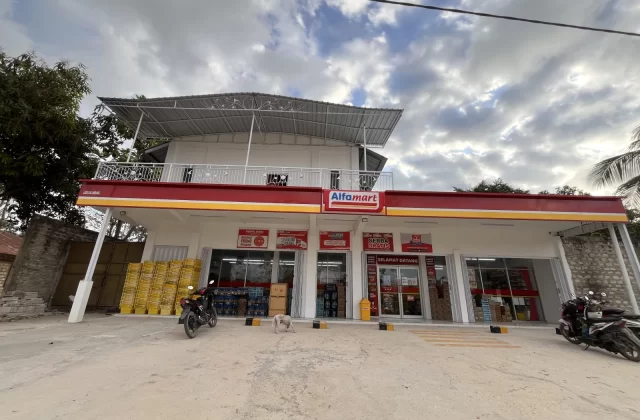 A modern white Alfamart minimarket with a red and yellow sign stands under a cloudy sky, with motorcycles and a small white dog outside.