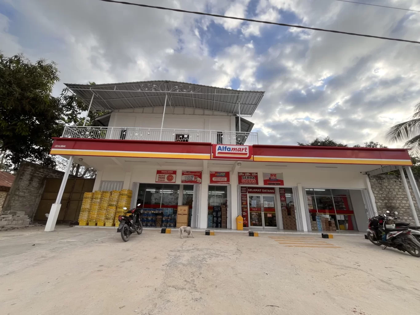 A modern white Alfamart minimarket with a red and yellow sign stands under a cloudy sky, with motorcycles and a small white dog outside.