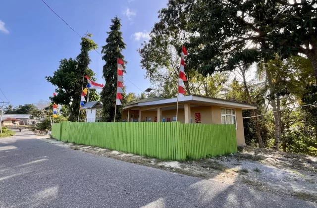 Matanyira Malaria Clinic building, light brown with a green fence, stands beside an asphalt road under a blue sky, adorned with Indonesian flags.