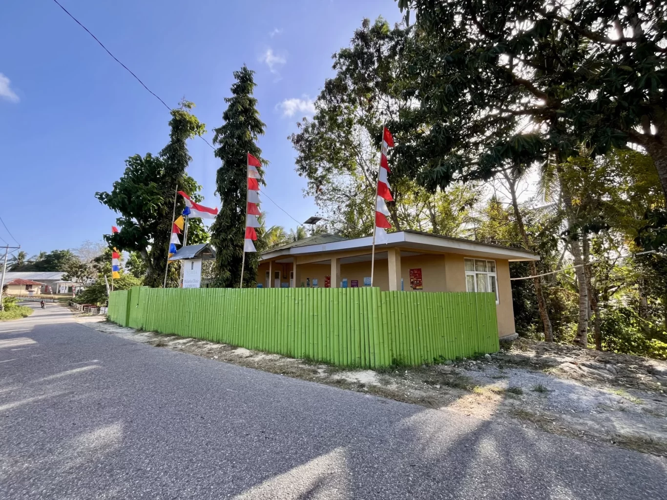 Matanyira Malaria Clinic building, light brown with a green fence, stands beside an asphalt road under a blue sky, adorned with Indonesian flags.