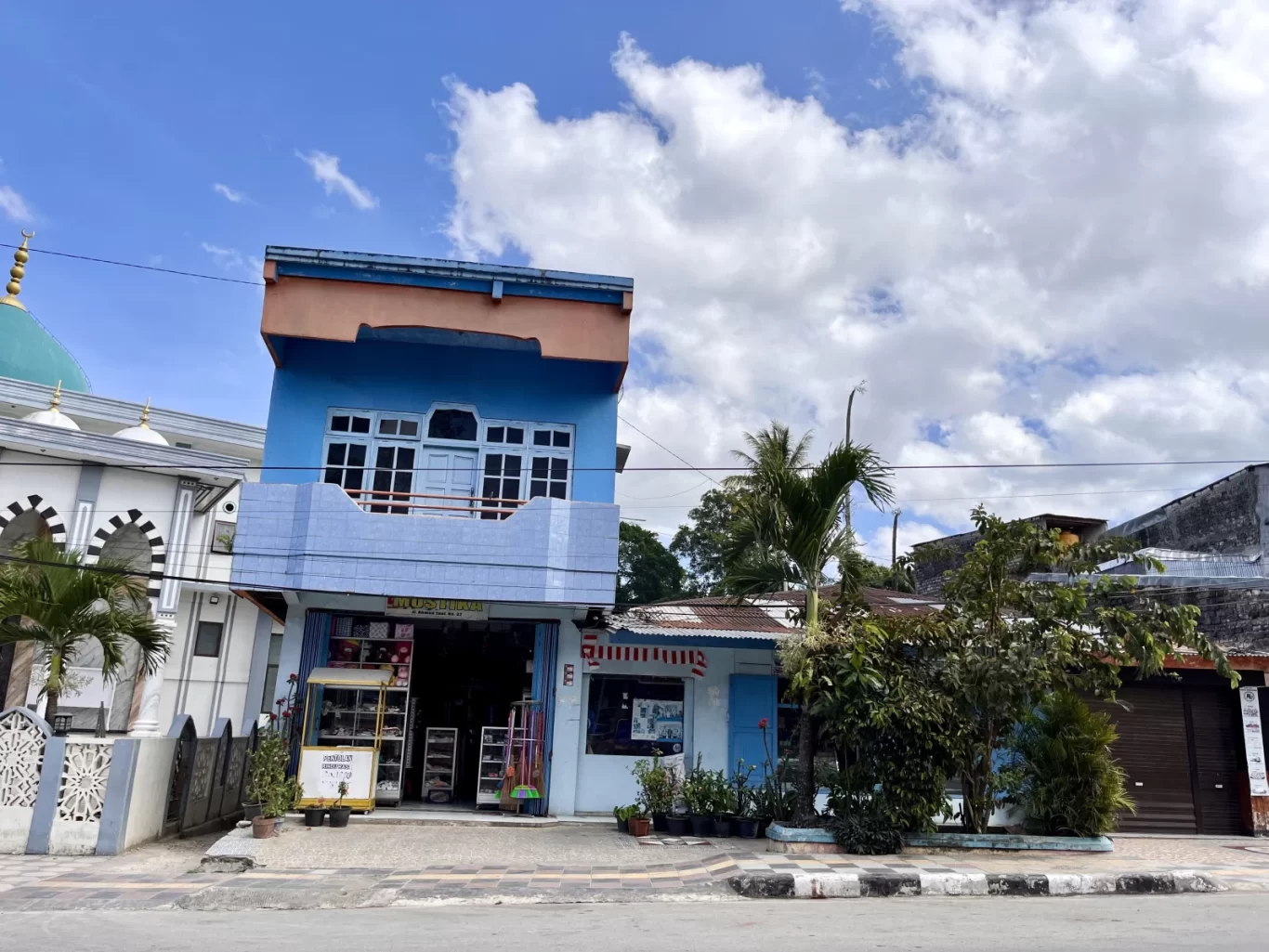 A blue two-story Mustika Store with an orange top stands on a street in Waikabubak, West Sumba, next to a mosque under a cloudy sky.