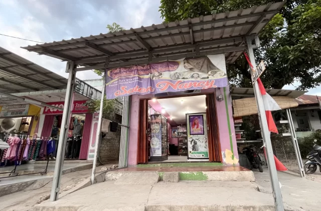 A small, pink-painted beauty salon named 'Salon Nancy' with a corrugated metal roof stands on a street in Tambolaka, Sumba, under a cloudy sky.