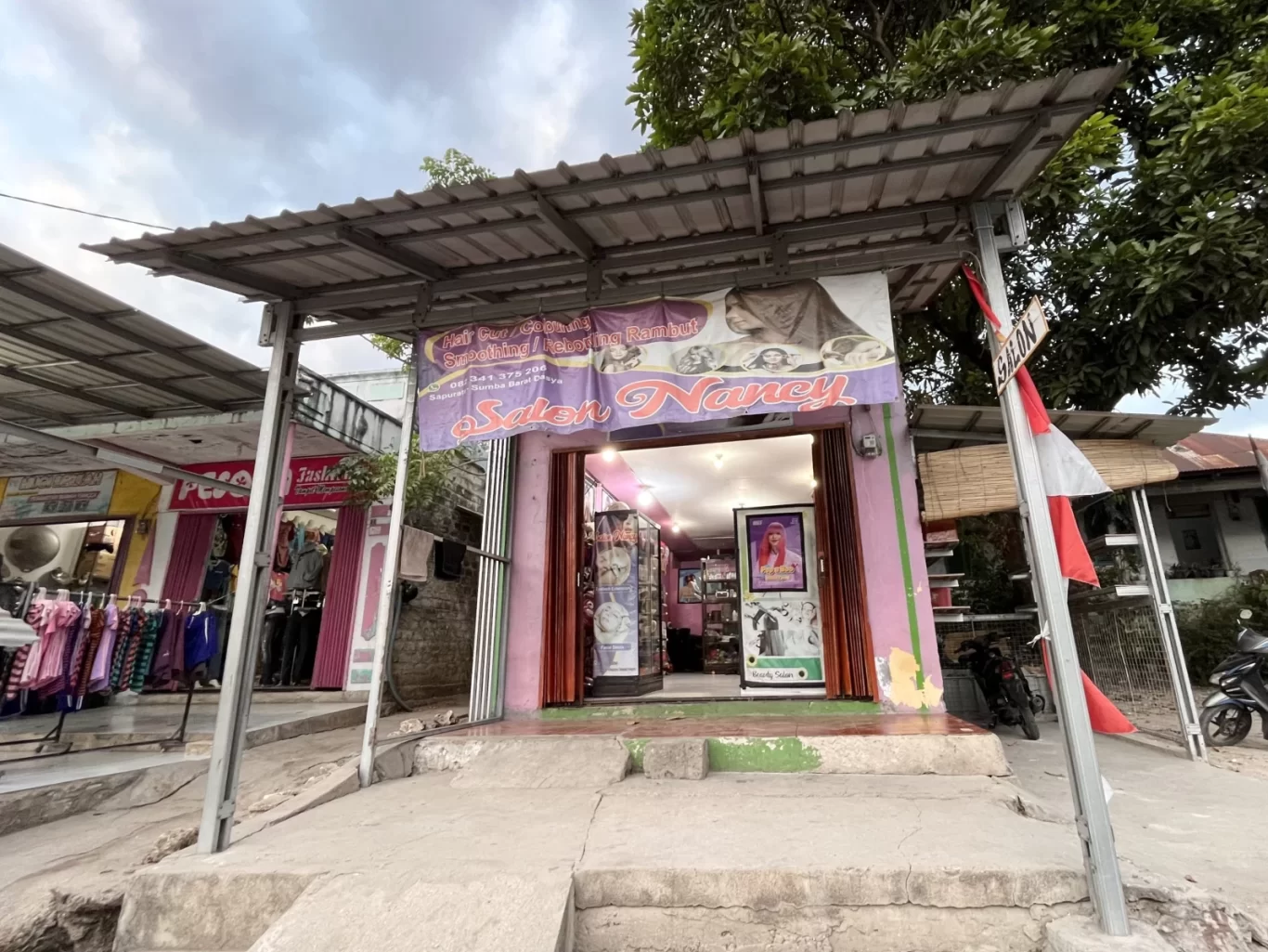 A small, pink-painted beauty salon named 'Salon Nancy' with a corrugated metal roof stands on a street in Tambolaka, Sumba, under a cloudy sky.
