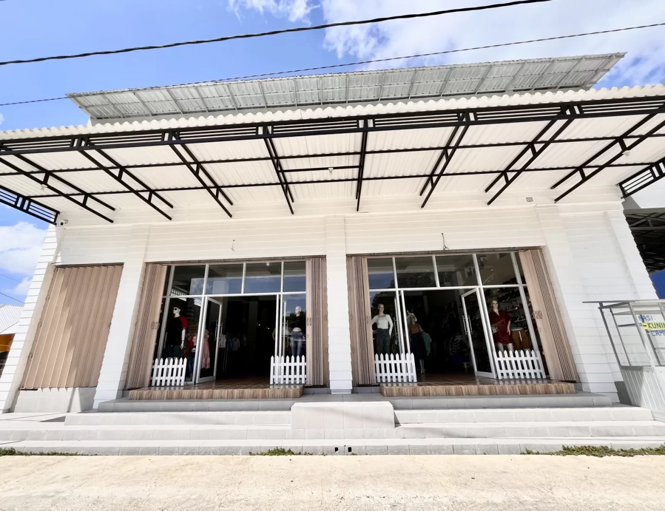 The white Oretz Boutique storefront in Waikabubak, West Sumba, features two glass entrances with mannequins displaying clothing under a black metal awning and blue sky.