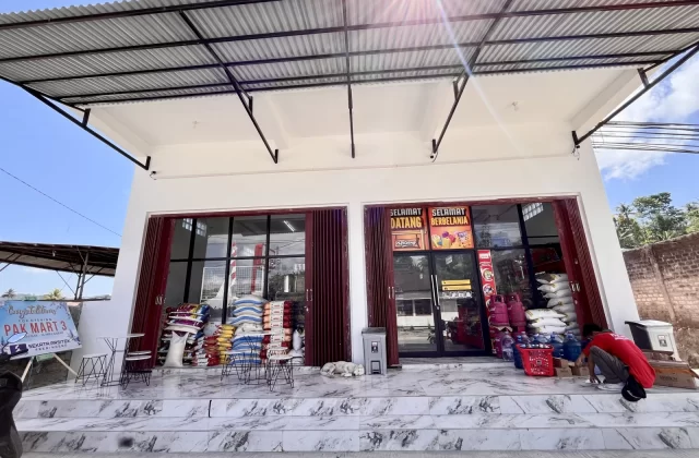 The Waikabubak Pak Mart minimarket features a white building with glass doors, stacked goods, a crouching person, and a sleeping dog under a bright blue sky.