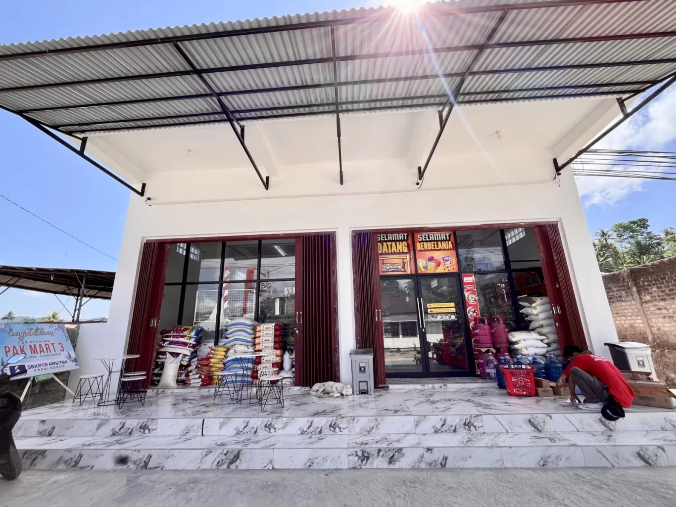 The Waikabubak Pak Mart minimarket features a white building with glass doors, stacked goods, a crouching person, and a sleeping dog under a bright blue sky.