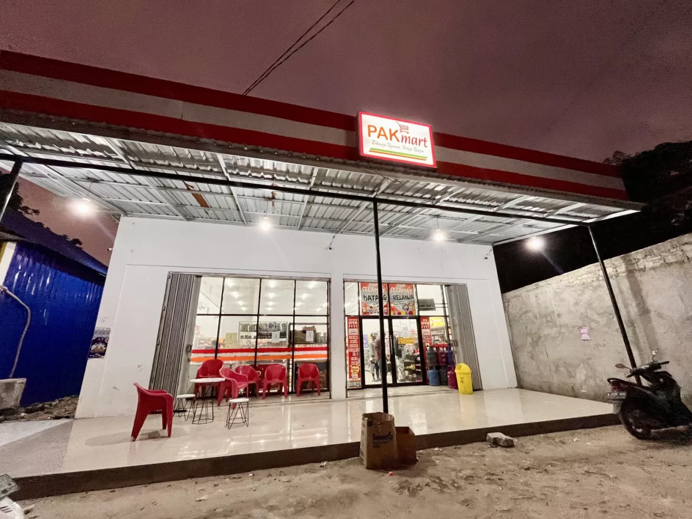 The brightly lit exterior of Waimangura Pak Mart minimarket at night, featuring a red and white sign, glass windows, and red outdoor seating.