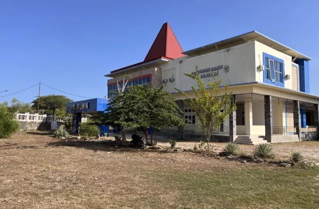 A large, modern Pratama Hospital building with a red roof and blue accents stands in a dry, grassy field under a clear blue sky in West Sumba.