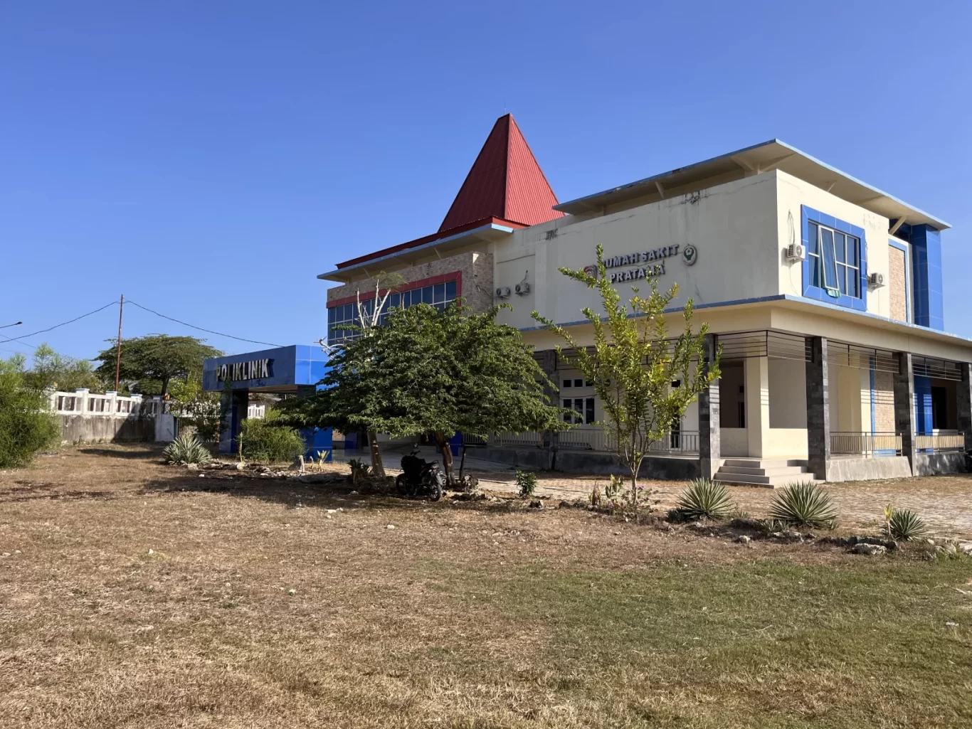 A large, modern Pratama Hospital building with a red roof and blue accents stands in a dry, grassy field under a clear blue sky in West Sumba.