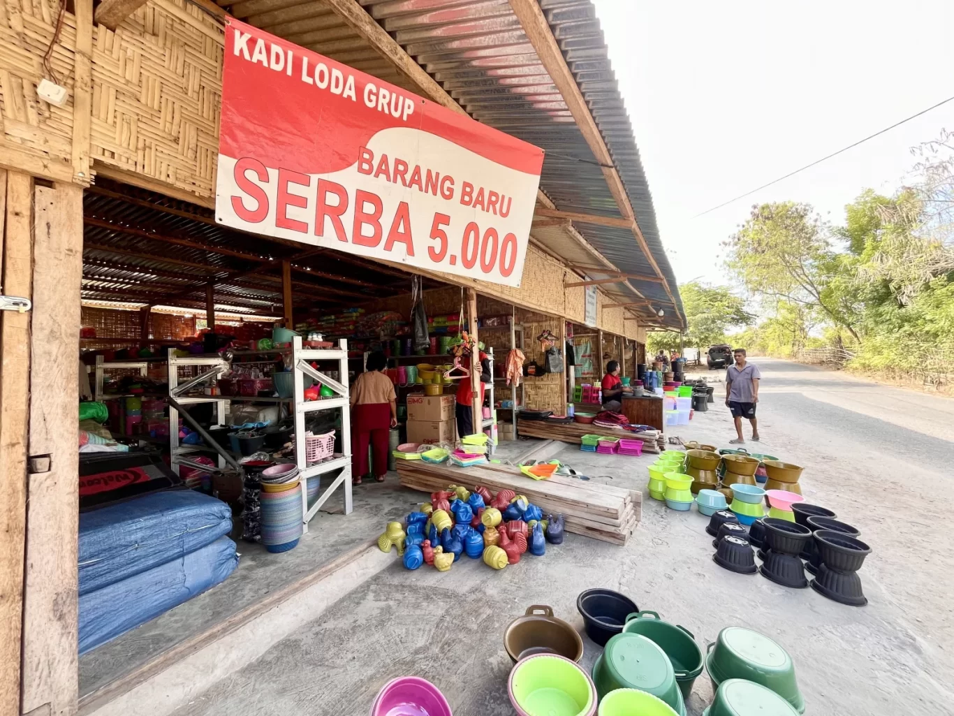 A vibrant roadside Serba 5.000 store in Tambolaka, Sumba, displays numerous colorful plastic household items, with customers inside and a man walking past.