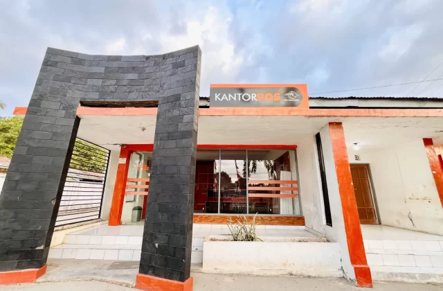 The Tambolaka Post Office building features white walls, orange accents, and a distinctive black curved entrance structure under a cloudy sky.