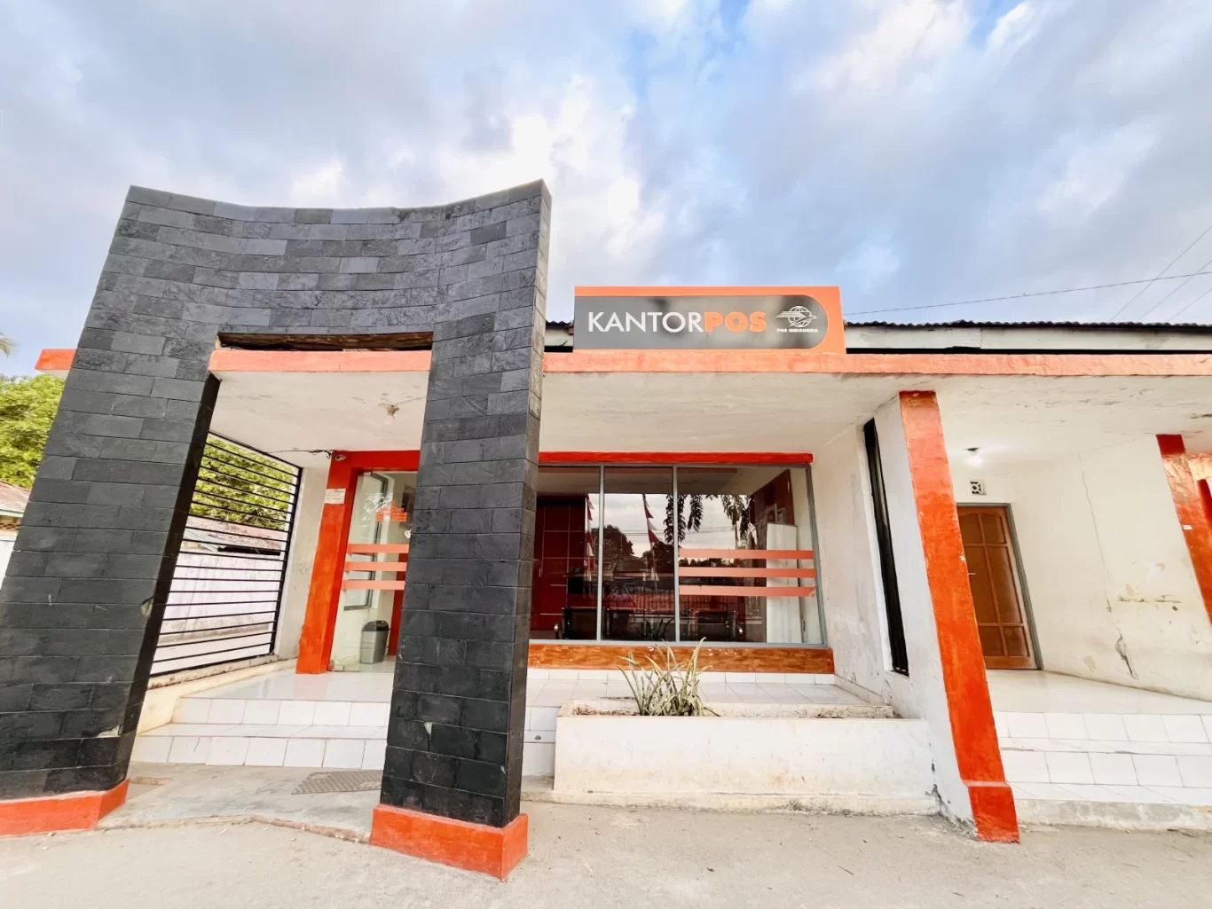 The Tambolaka Post Office building features white walls, orange accents, and a distinctive black curved entrance structure under a cloudy sky.