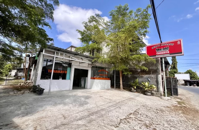 The exterior of Tanakatto Restaurant in Waikabubak features a white building with glass windows, a red sign, and a concrete parking area under a blue sky with trees.