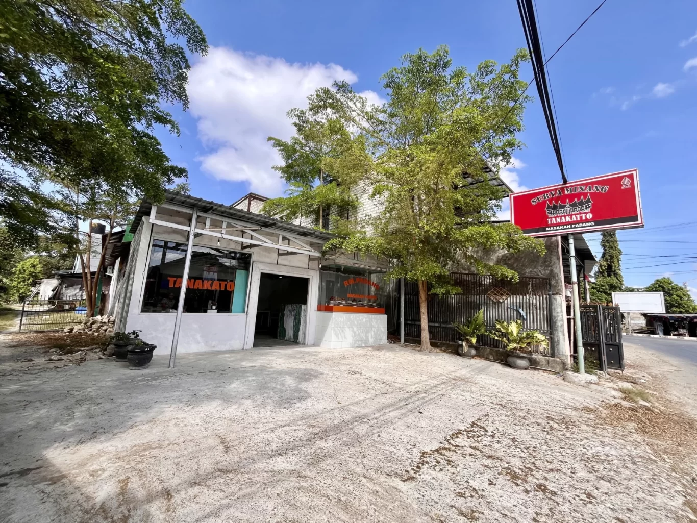The exterior of Tanakatto Restaurant in Waikabubak features a white building with glass windows, a red sign, and a concrete parking area under a blue sky with trees.