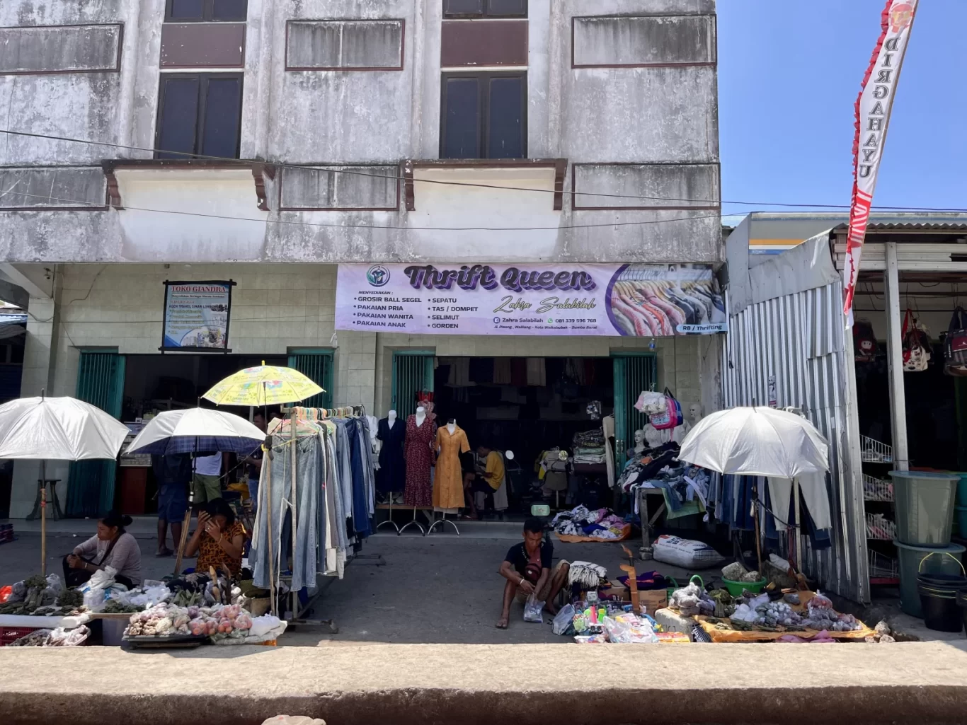 A weathered concrete building houses "Thrift Queen" with a banner, while street vendors sell clothing and produce under umbrellas on a sunny day in Waikabubak.