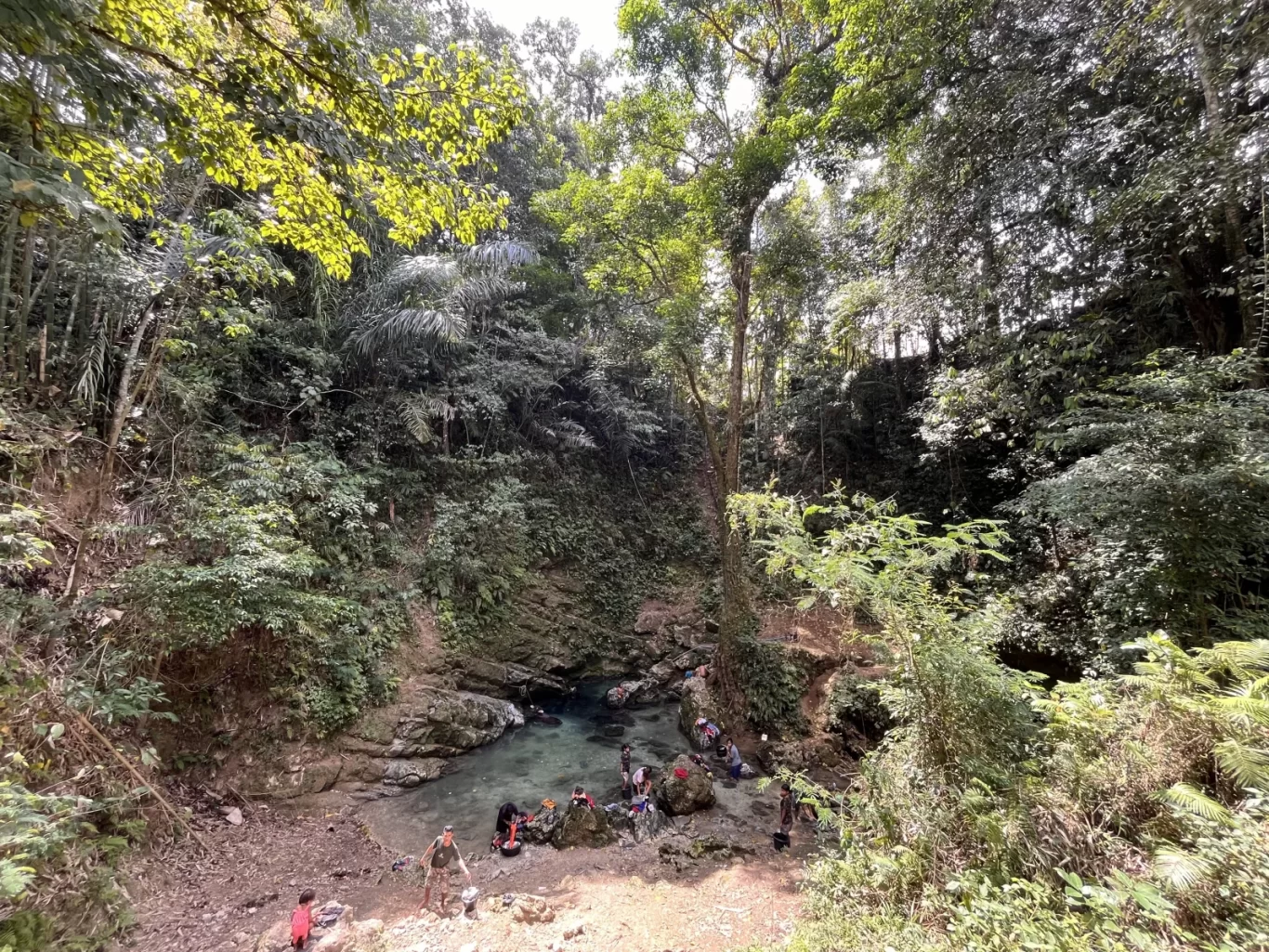 A group of people gathers around a clear blue natural spring pool surrounded by dense green jungle foliage under dappled sunlight.