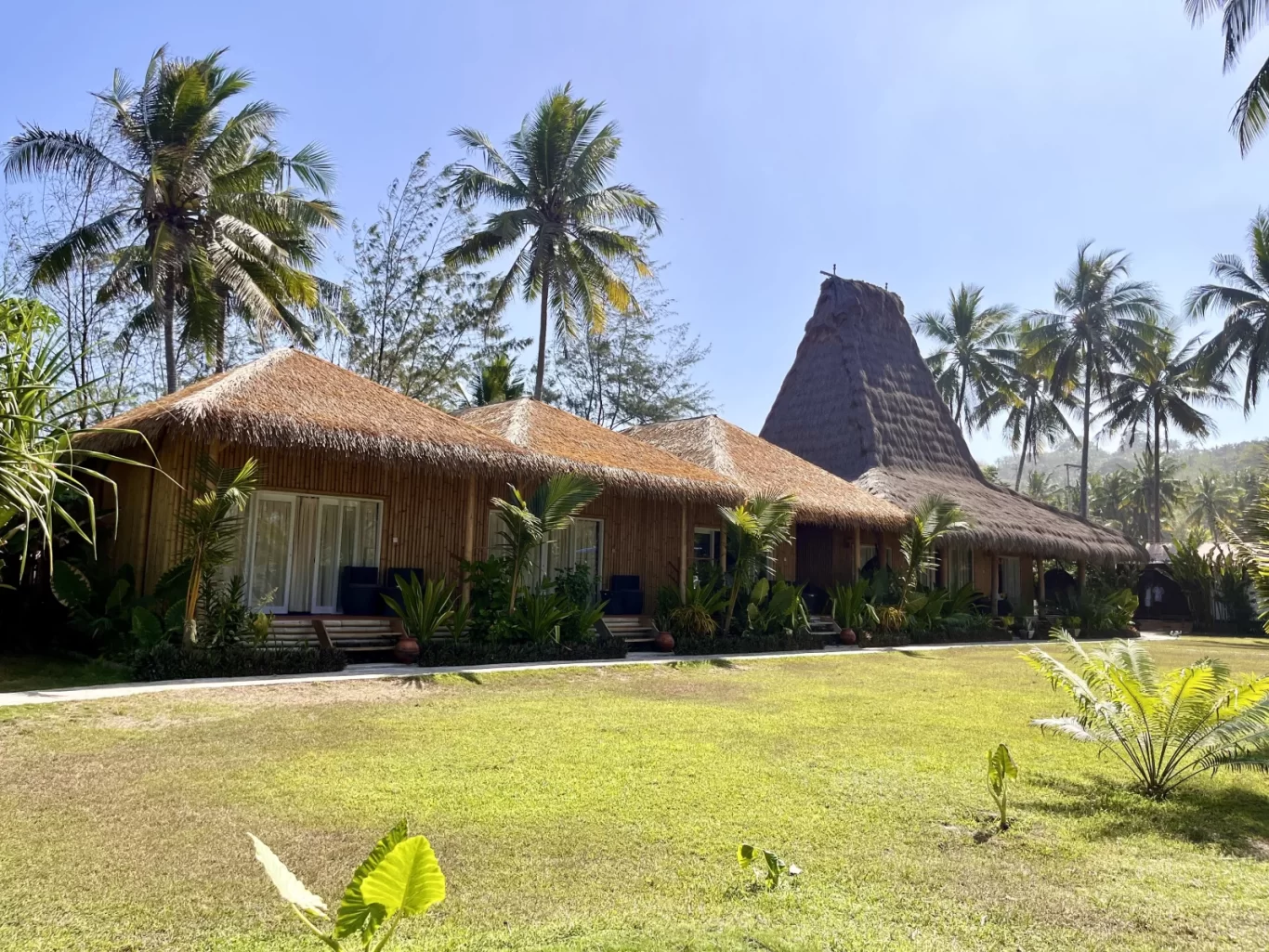 Traditional Sumbanese-style bungalows and a tall conical-roofed building stand on a green lawn surrounded by palm trees under a clear blue sky.