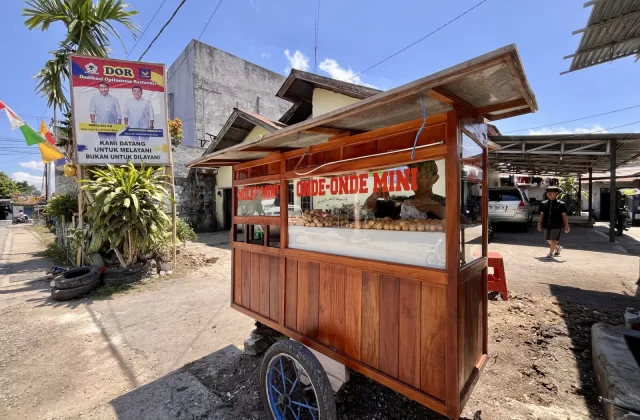 A wooden Onde-Onde Mini food stall stands on a dirt path, displaying numerous round pastries under a bright blue sky in Waikabubak, Sumba. A political banner and palm trees are visible nearby.