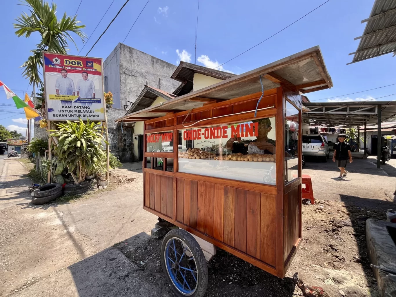 A wooden Onde-Onde Mini food stall stands on a dirt path, displaying numerous round pastries under a bright blue sky in Waikabubak, Sumba. A political banner and palm trees are visible nearby.