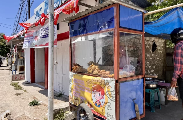 A blue and brown street food stall, DD Azhar 75, displays fried Molen and other Indonesian snacks on a sunny street in Waikabubak, Sumba.