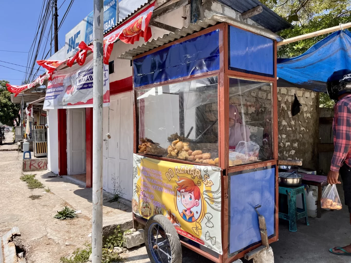 A blue and brown street food stall, DD Azhar 75, displays fried Molen and other Indonesian snacks on a sunny street in Waikabubak, Sumba.