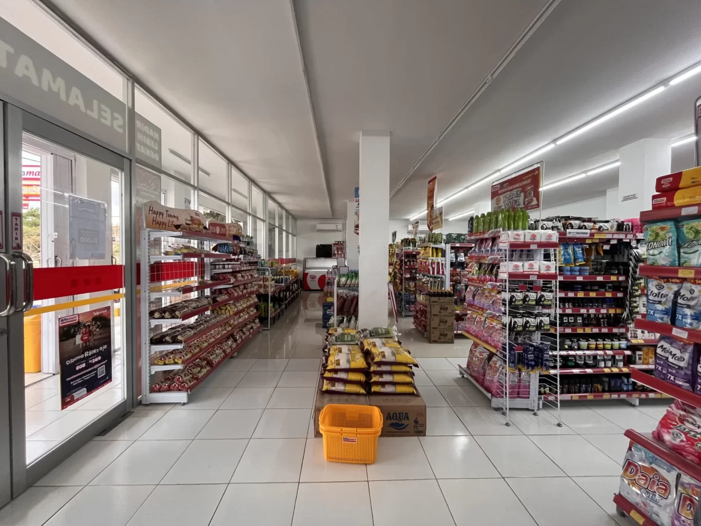 The brightly lit interior of an Alfamart minimarket in Loura, Sumba, features white tiled floors, multiple aisles stocked with diverse products, and glass entrance doors on the left.