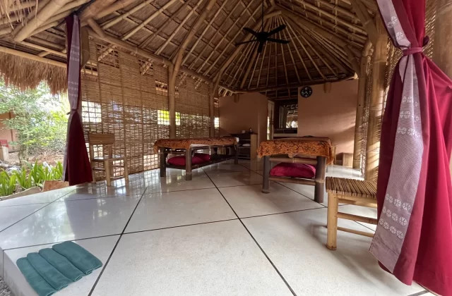 A serene open-air spa pavilion in Sumba features two massage beds with red cushions, a thatched roof, bamboo walls, white tiled floor, and red patterned curtains.