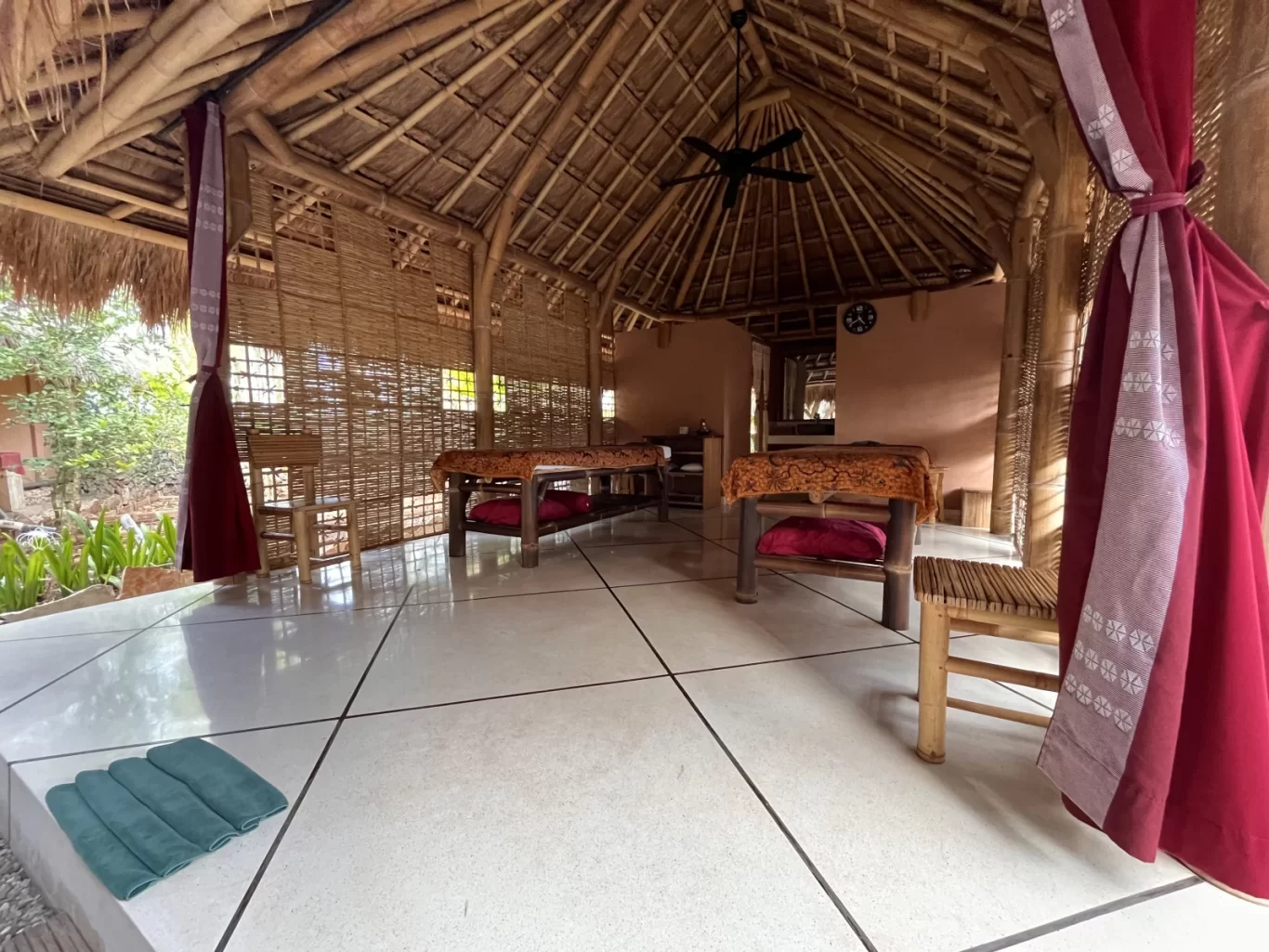 A serene open-air spa pavilion in Sumba features two massage beds with red cushions, a thatched roof, bamboo walls, white tiled floor, and red patterned curtains.