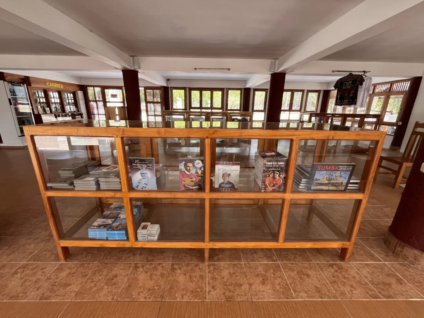 Interior of Rumah Budaya Sumba Souvenir Store, showing a wooden display case with books and items, surrounded by windows and more displays.